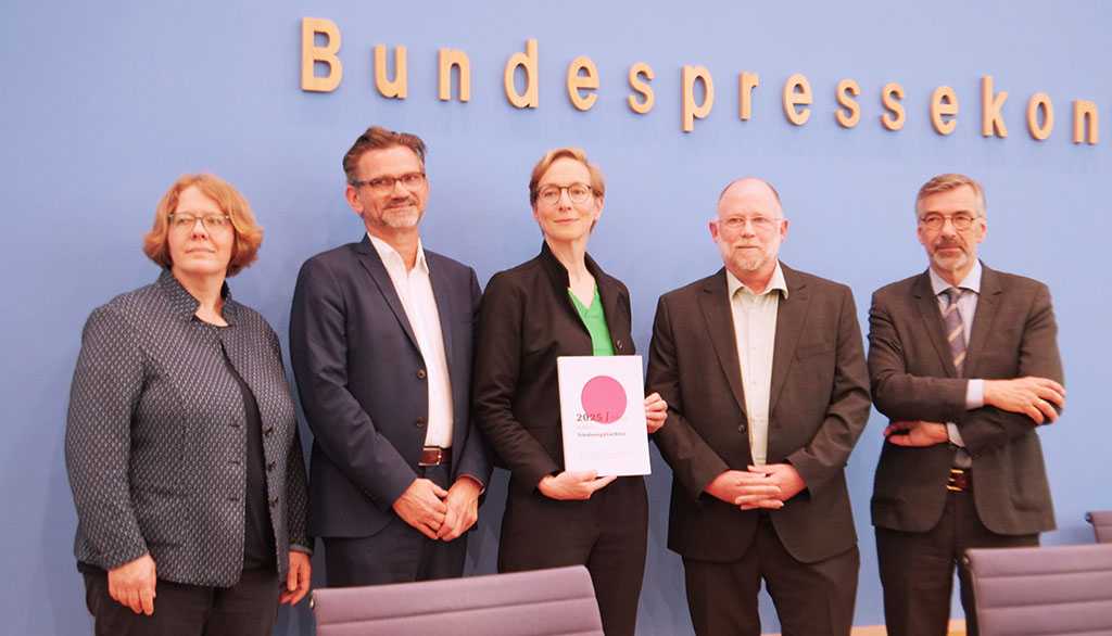 Fünf Personen vor dem Schriftzug Bundespresseko, eine Frau in der Mitte hält das Friedensgutachten 2025 in der Hand.