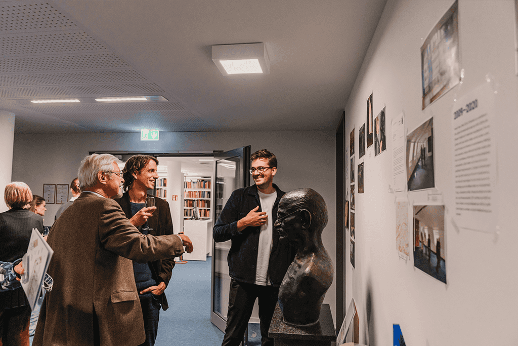 Eine Gruppe von Männern, die eine Wand mit aufgehängten Fotos und Texten betrachten.