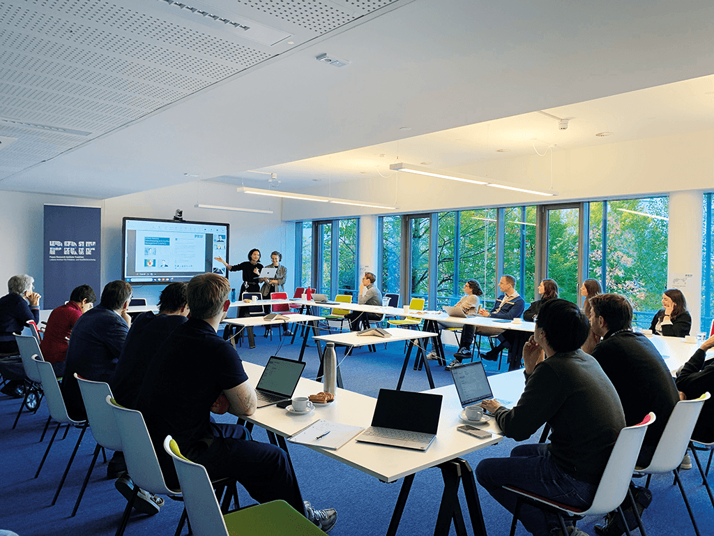 Meetingraum mit vielen Menschen, zwei Personen stehen an einem Screen.