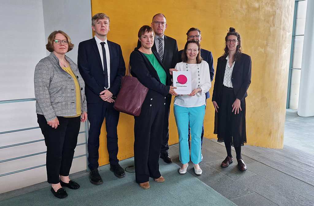 Eine Gruppe von Personen steht vor einer gelben Wand, eine Frau in der Mitte überreicht einer Frau daneben das Friedensgutachten 2025.